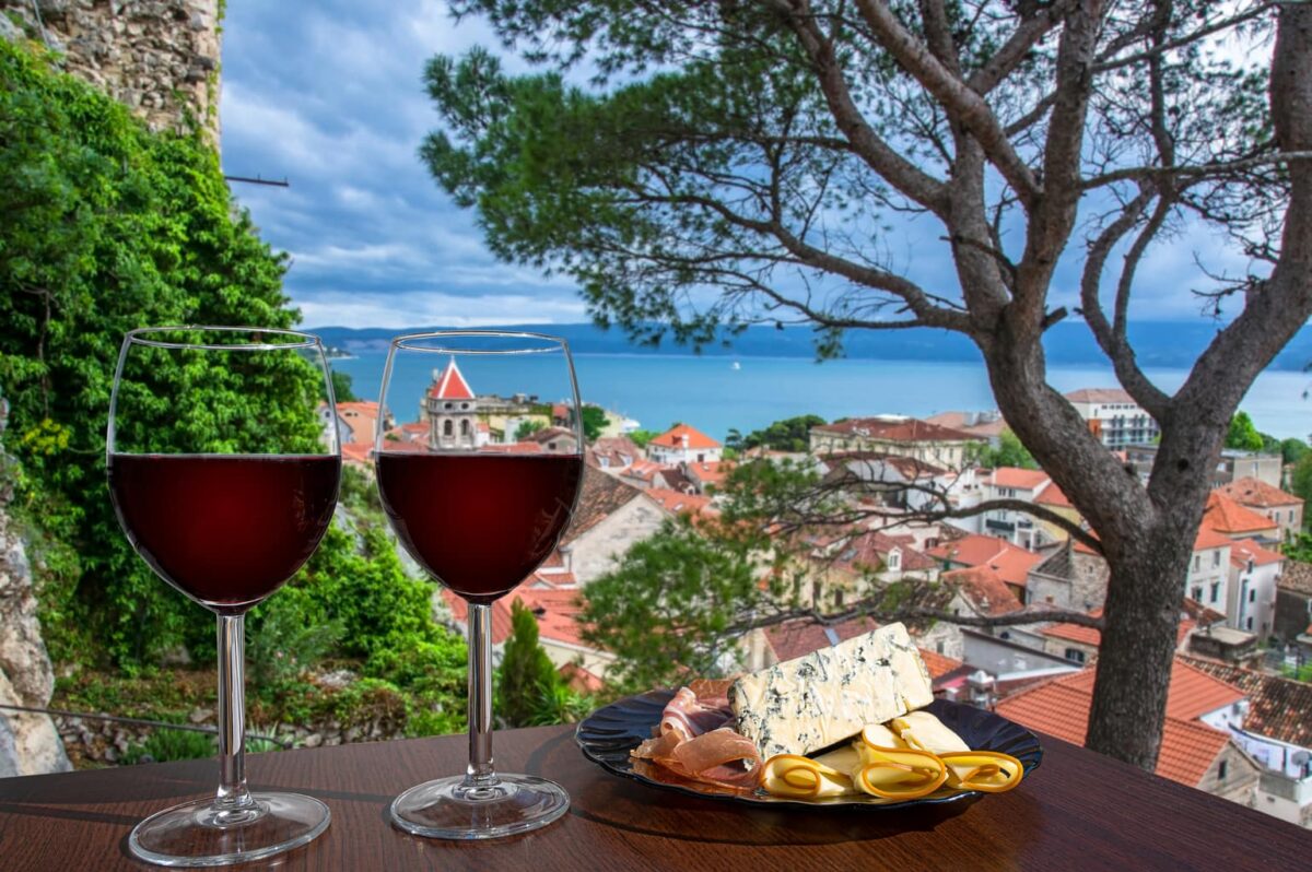 Two glasses of red wine with charcuterie assortment with view of Croatian town Omis with red roofs and blue sea. Glass of red wine with different snacks - plate with ham, sliced, blue cheese.