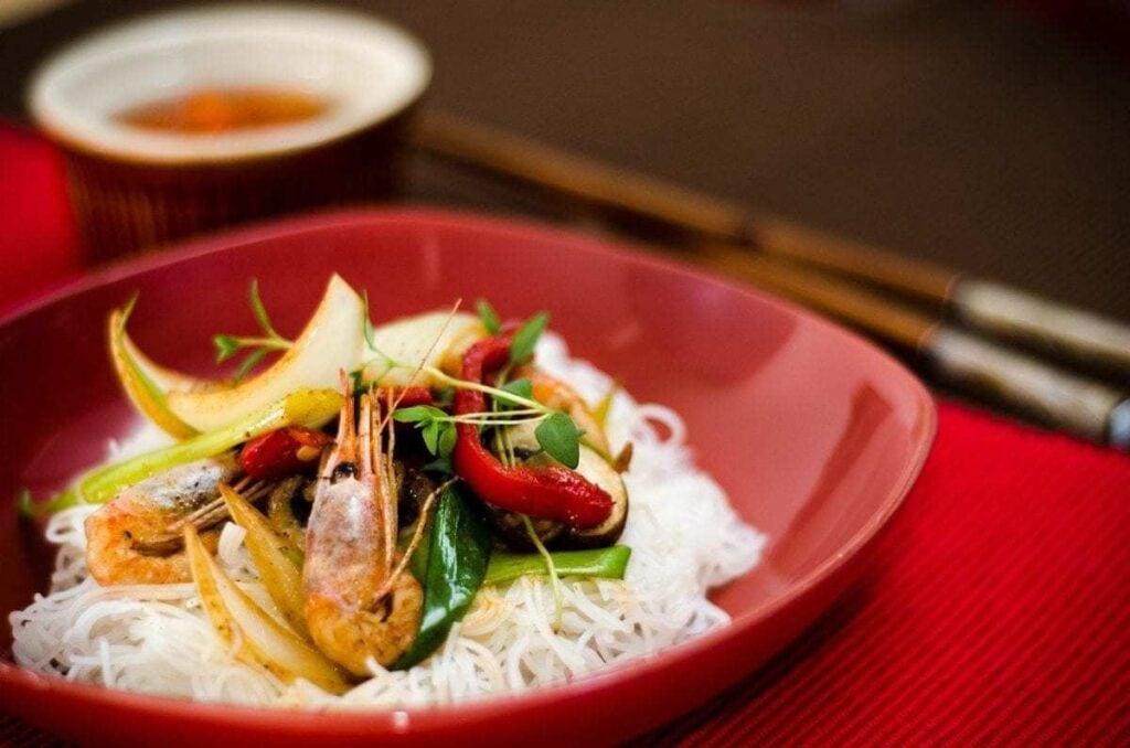 UNESCO Intangible Cultural Heritage a dish of rice noodles in a red bowl, with a prawn, red pepper slices and pea shoots decorating the dish. There are chopsticks beside the bowl which sits on a red placemat