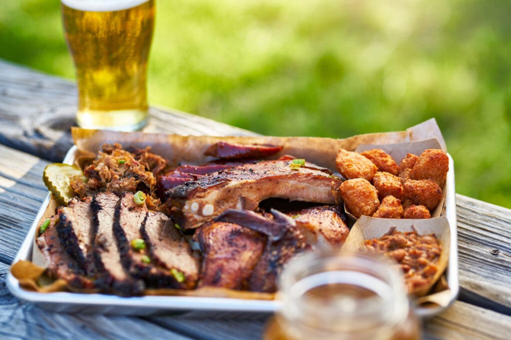 A platter of BBQ, including nuggets, ribs, and brisket all loaded onto a silver tray and sitting on a picnic bench with 2 beers - Weird American Food