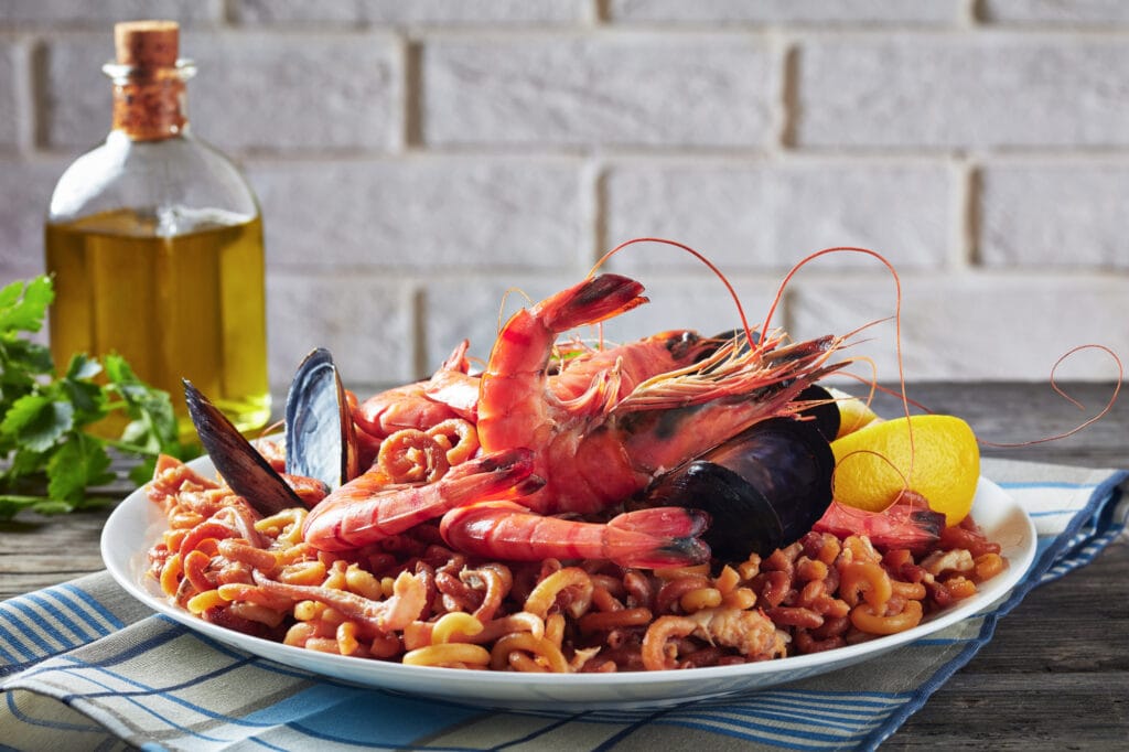 A plate of Spanish seafood pasta with shrimp and mussels, accompanied by a bottle of olive oil and lemon on a wooden table.