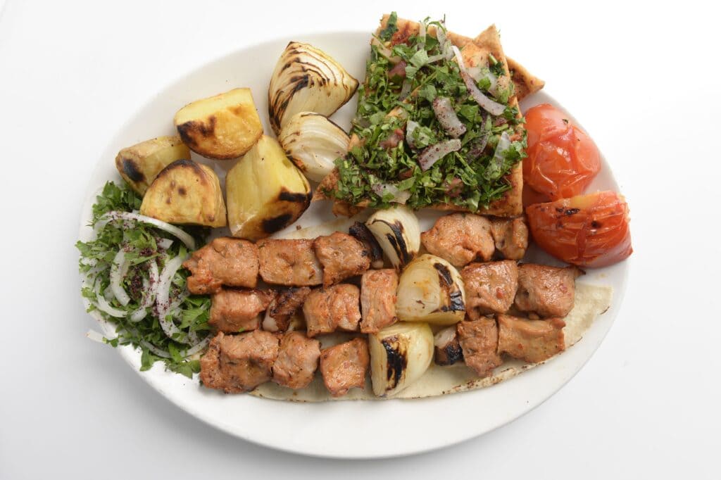 Lebanese Food – a white plate with pita bread triangles covered in onions nad herbs, cubed chicken from the BBQ, roasted onions and tomatoes