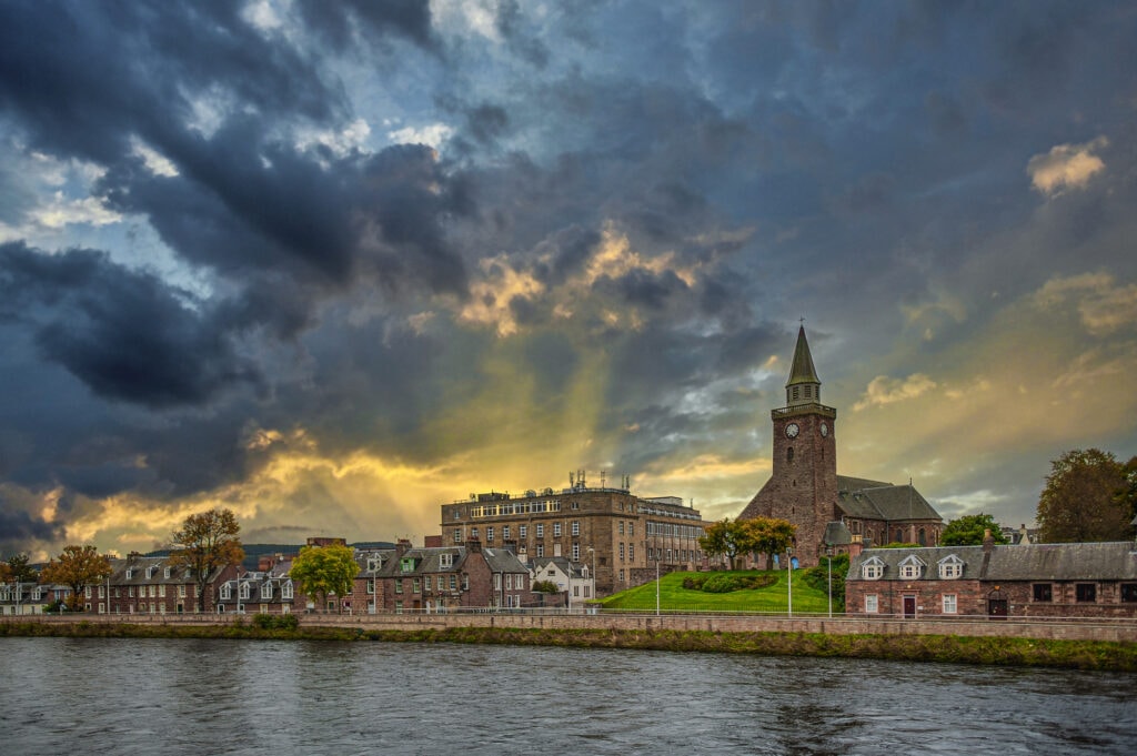 Things to do in Inverness, Scotland a gorgeous view of Inverness across the river, the sky is dark and cloudy with the sun breaking through into golden rays of light.