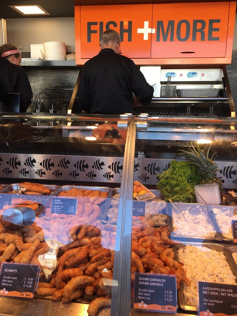 What to eat in Amsterdam a kibbeling booth which serves up deep-fried pieces of fish and scampi. The sign in orange says fish+more and the man is serving up quick to go kibbeling snacks.