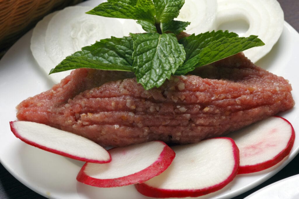 Lebanese food - a plate of kibbeh which is on a white platter, the meat is raw and shaped like a football and surrounded by a soft cream cheese and radish slices with a mint leaf on top