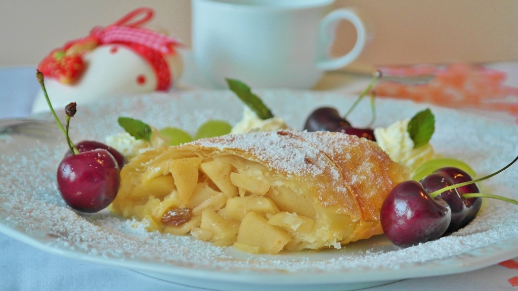 German Foods a plate with an apple strudel in the centre with apples and raisins spilling out of the strudel which is decorated with cherries and powered sugar