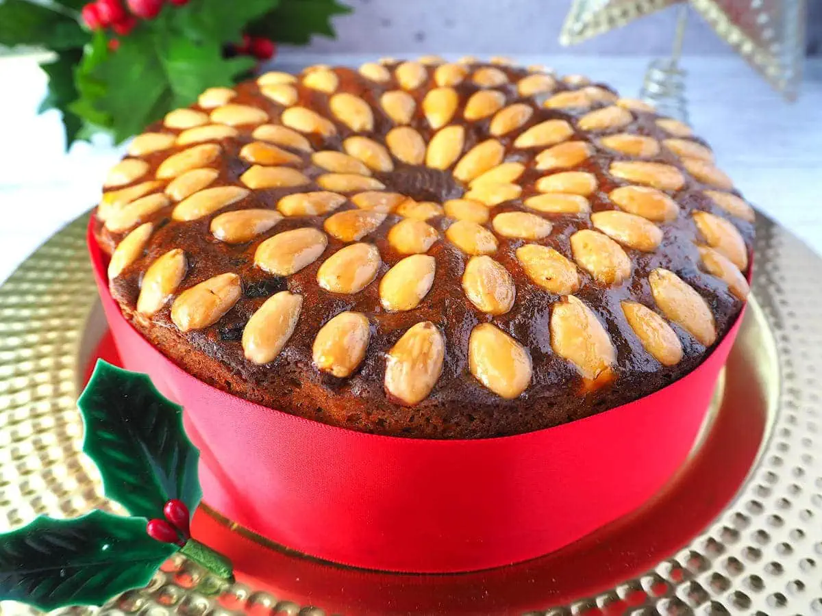 A round fruit cake filled with an assortment of dried fruits and lots of booze with a red ribbon around it and whole almonds decorating the top