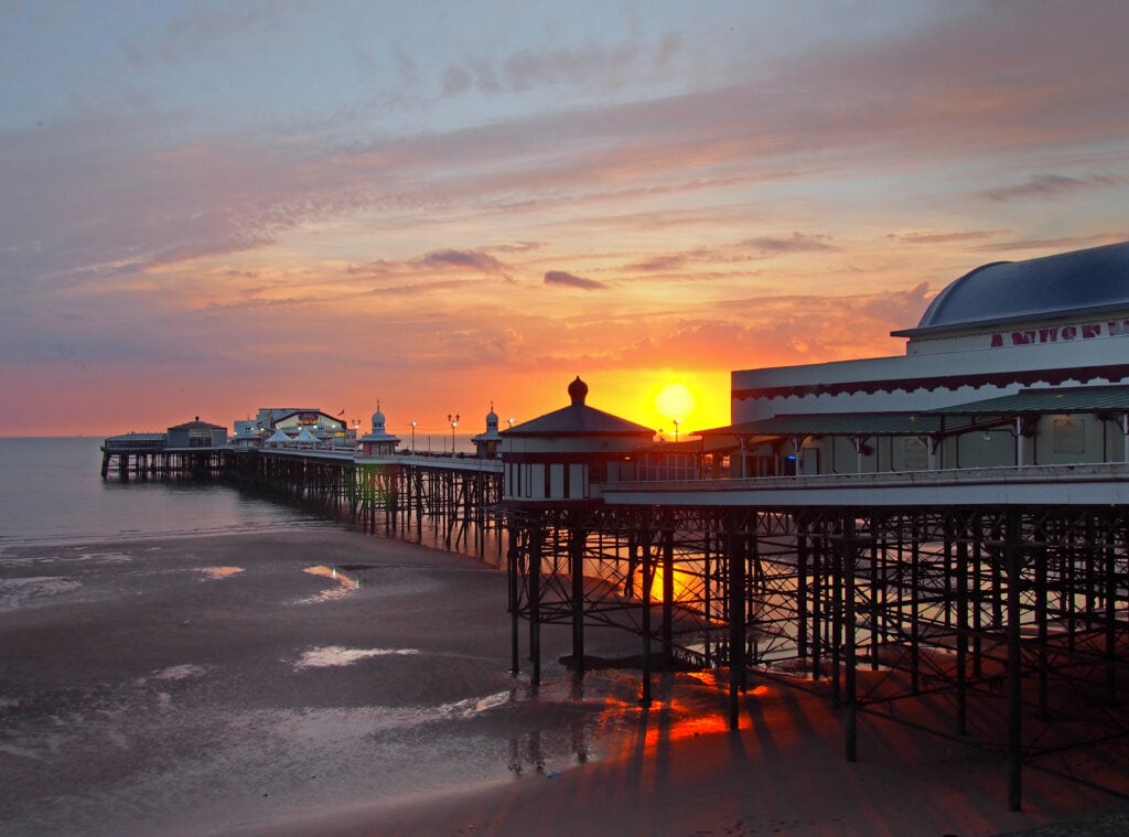 the sun setting over the historic north pier in blackpool with glowing light reflected on the beach and colourful twilight sky