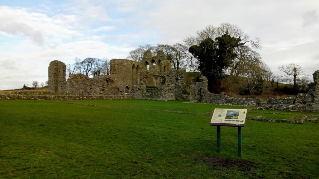 The crumbling ruins of Inch Abbey where Game of Thrones the War of 5 Kings was filmed. The ruins are a mainly stone walls with some high window arches left and an information board in front of the ruins. 