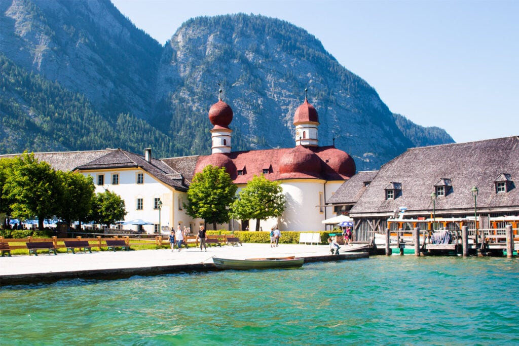 View of Konigssee Lake - Germany Travel Guide. Mountains rise up behind a church with onion domes and the dock beside the beautiful blue lake.