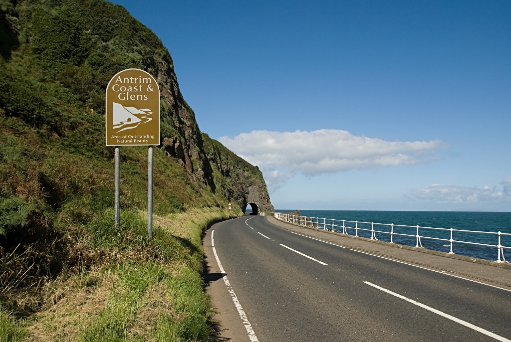 Sign on the Antrim road that leads to the Glens of Antrim. To the left is the north sea and up ahead is an arch cut out of the stone cliff that covers the road