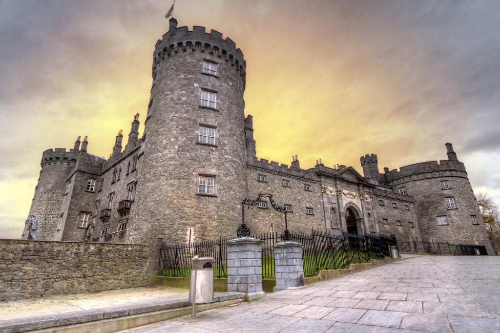 The most beautiful castle in Ireland Kilkenny is made from grey stone with crenellated turrets on all four corners a small patch of green lawn in front and a great arched doorway where you enter