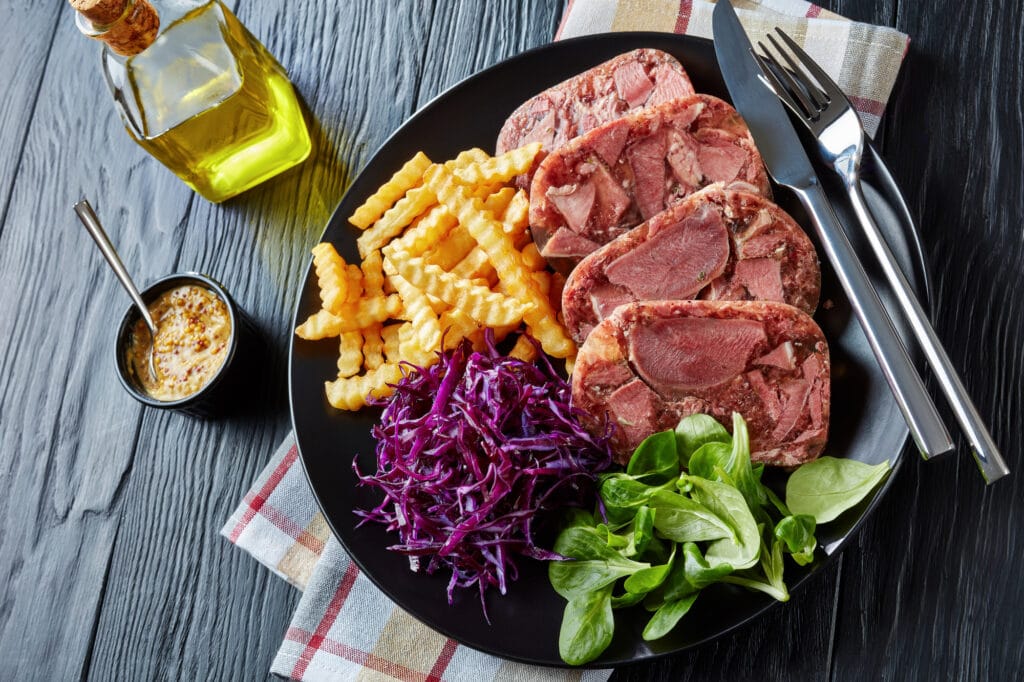 sliced beef tongue aspic served with french fries, green leaves and red cabbage salad on a black plate on a wooden table with mustard in a bowl, view from above, close-up