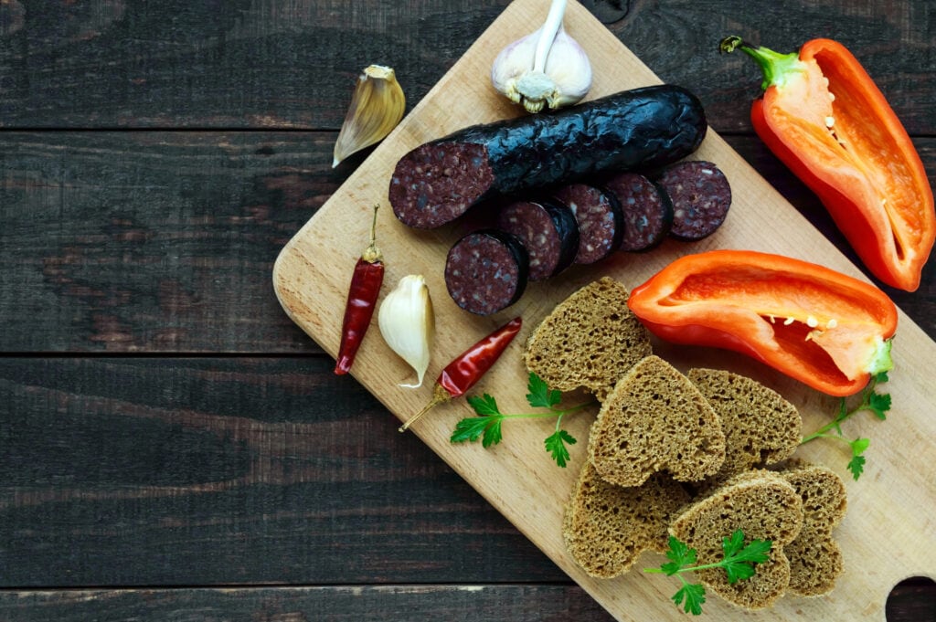 Morcillo (Spanish black pudding, blood sausage), cutting slices, black rye bread in a heart shape, pepper, garlic on a wooden board. The top view on a dark background. A festive meal on Christmas, Easter - types of sausage around the world