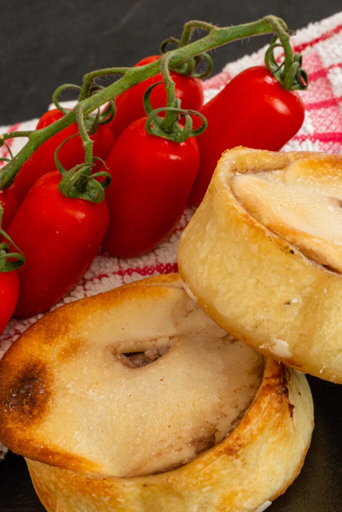 Two round Scotch Pies just out of the oven and served on a black slate serving plate and cherry vine tomatoes on the side..