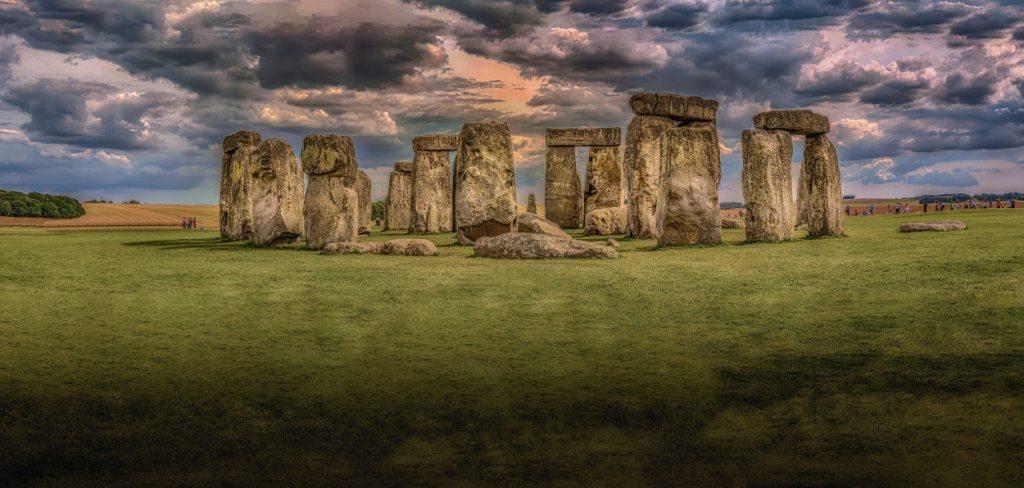 The famous stonehenge with a purple and pink coloured sky above then there are tiny people wandering the stones and the grass before and around the stones is a dull gray green
