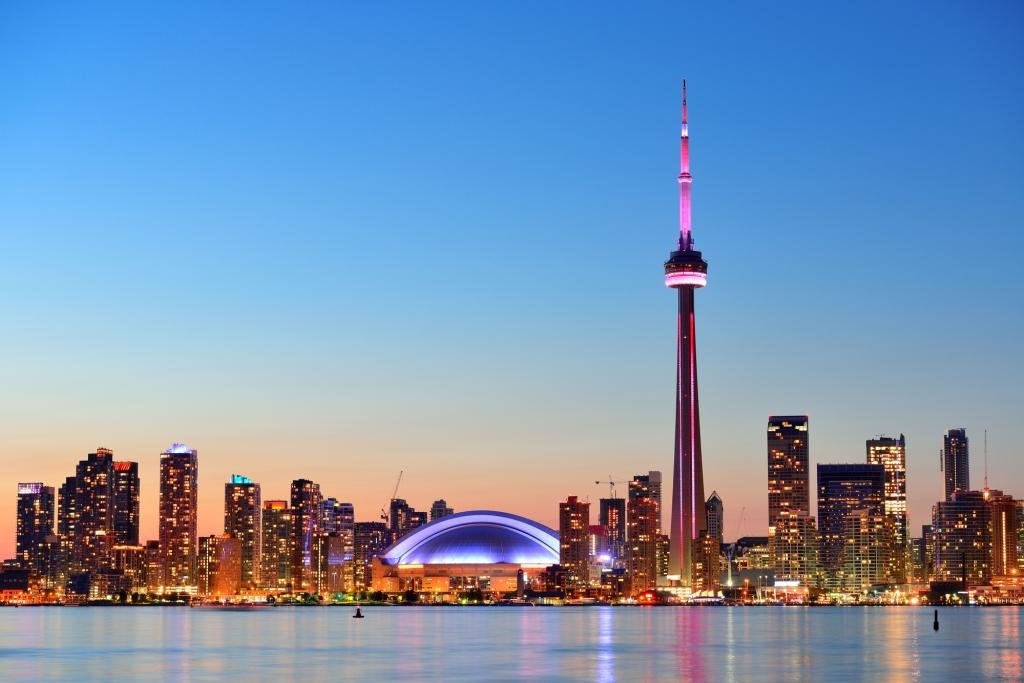 The skyline of Toronto at twilight with a pink and lavender sky, the CN tower and the lights of the high rises around the lake