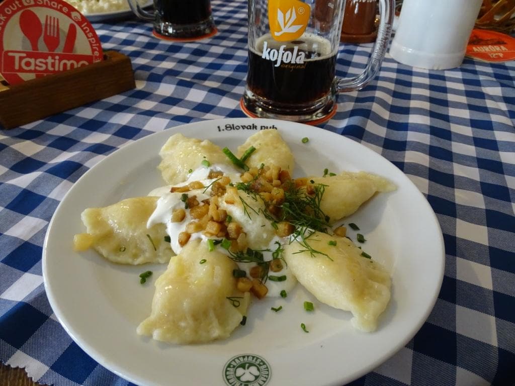  bryndzové pirohy, a delicious type of potato dumpling stuffed with the most popular ingredient in Slovakia: goat’s cheese. Shaped in half moon dough these are serve on a white plate and topped with sour cream and dill