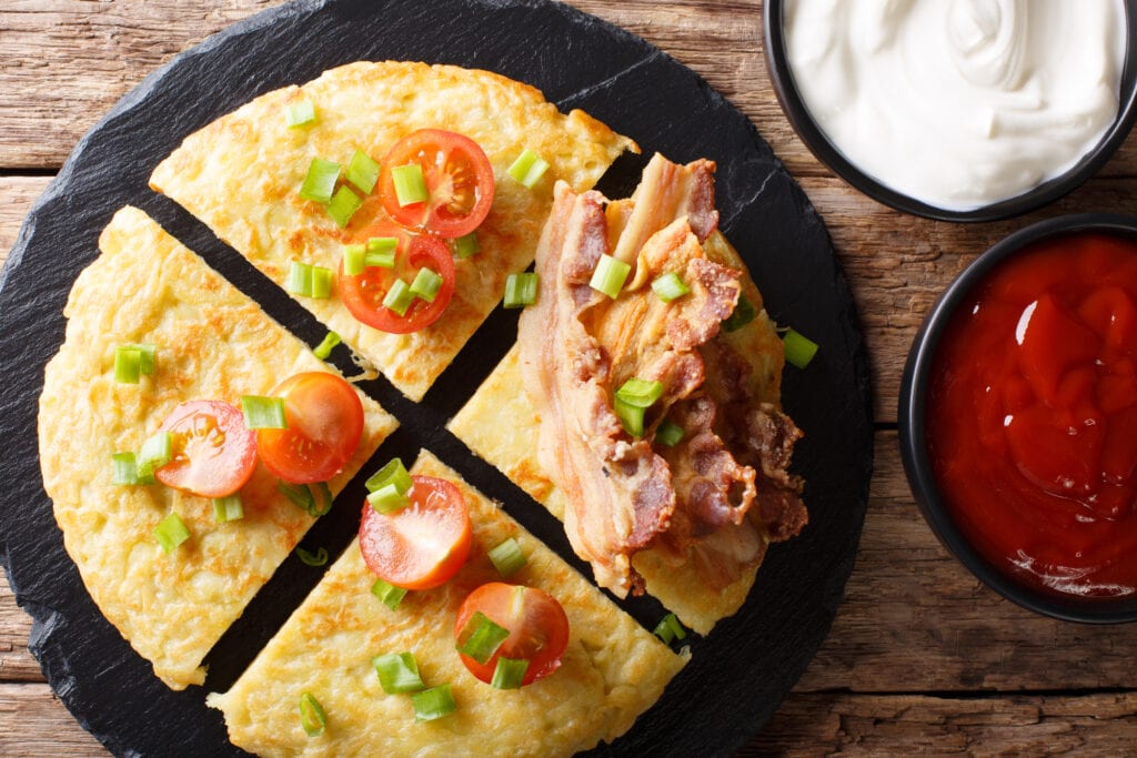 Potato boxty in a dark black griddle. The boxty is decorated with bacon tomatoes and onions. It is a lovely crispy brown shade