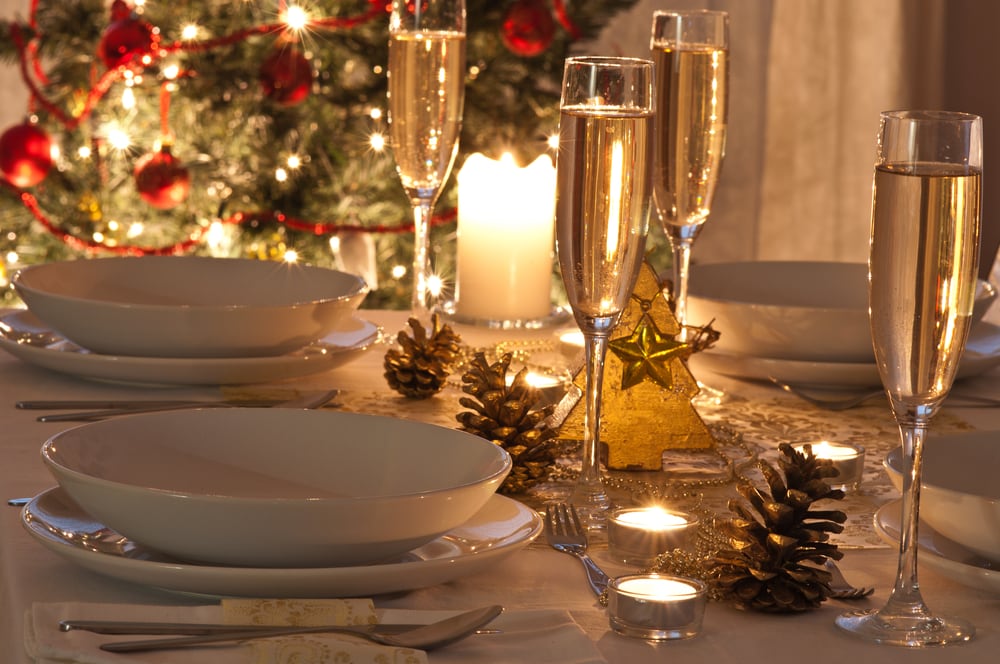 A Christmas table set with champagne glasses and white plates, decorated with golden pine cones and a Christmas tree in the background glowing with twinkly lights and red baubles