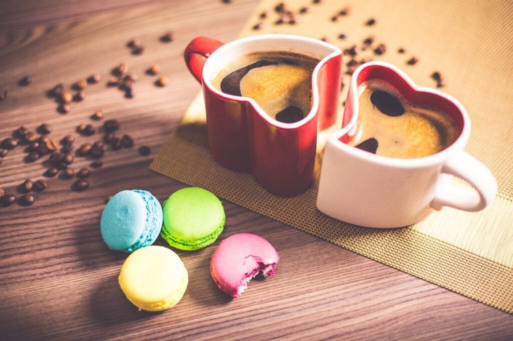 The History of Macarons - 4 macarons site on the table they are tiny and coloured pink, blue, green and white. There are 2 hear shaped coffee mugs full of espresso the mugs are red and white and there are coffee beans on the table