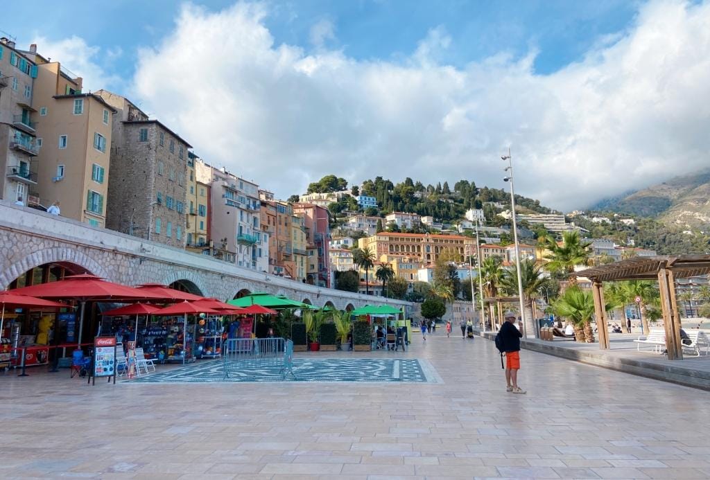 The seafront promenade in Menton France on the left are a series of bars and cafes with green and red sunshade umbrella, above those are a series of old apartment buildings up a hill with views of the promenade.