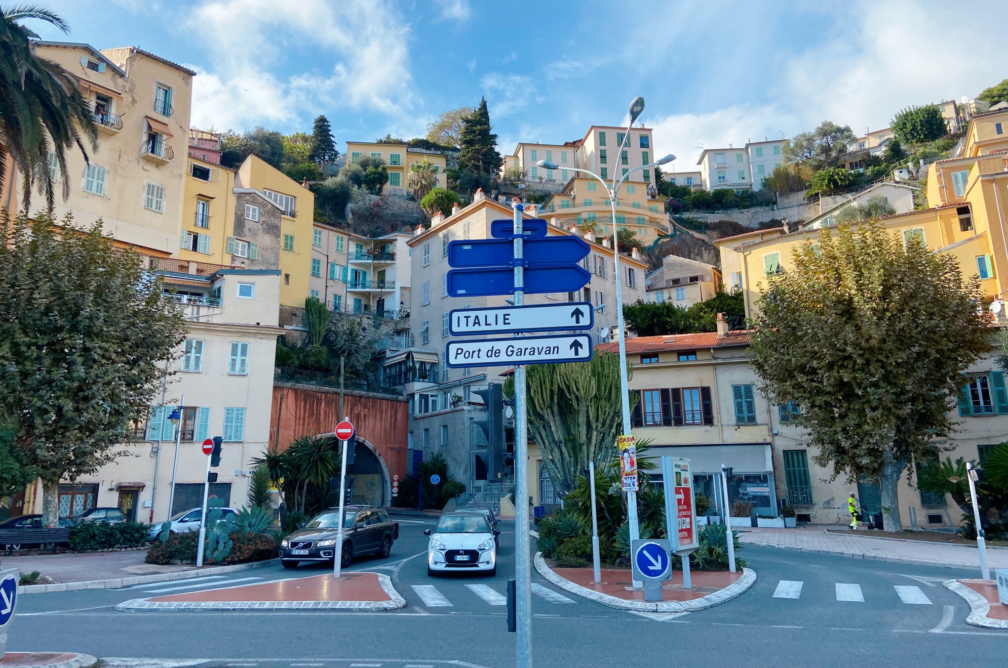 A sign in France detailing the area of Menton France. The road has a roundabout with several cars on it and the houses climb up a hill