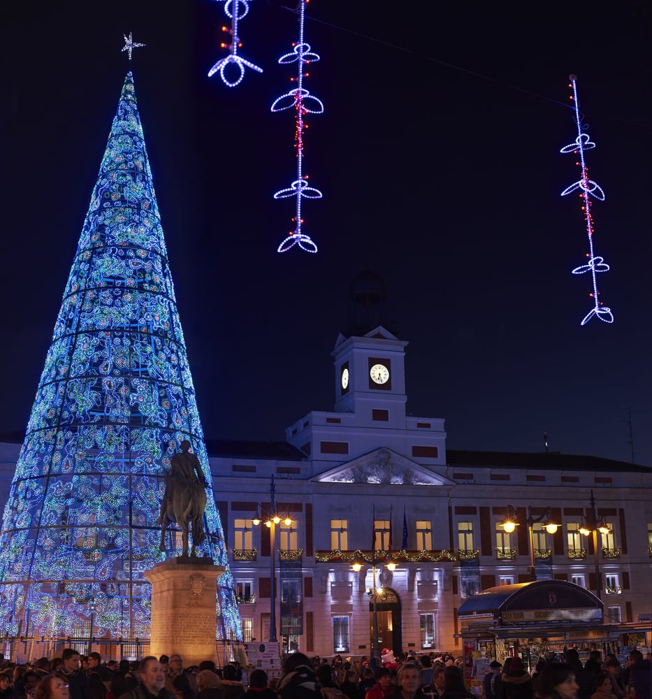 Puerta del Sol square at nightfall is illuminated by Christmas lights and a shiny Christmas tree with Real Casa de Correos in the background.
