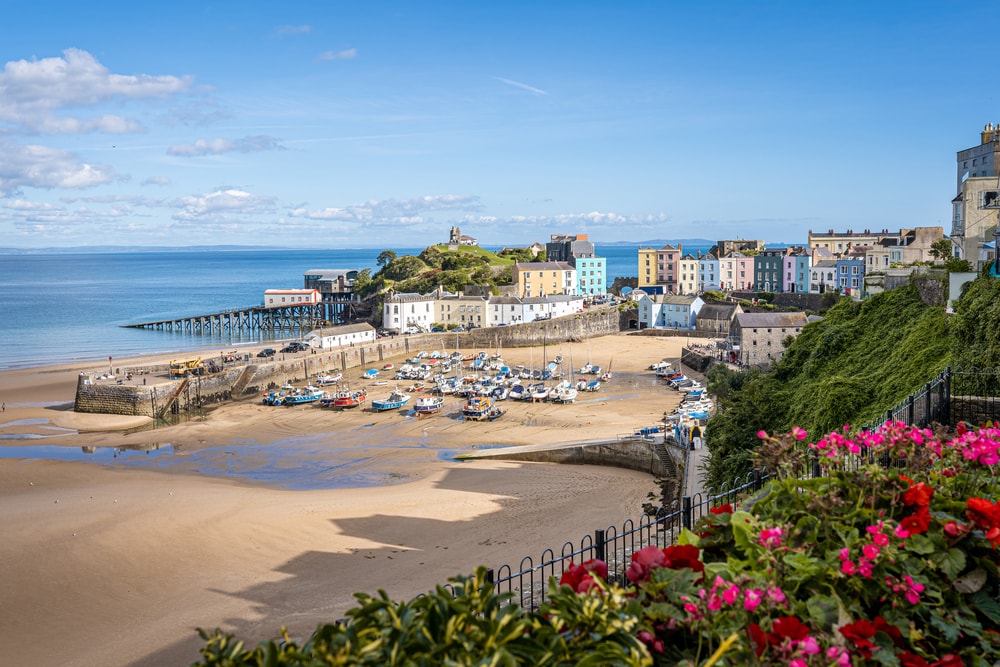 A view of Tenby harbour in Wales with the town sitting just above a cream coloured sandy beach. The sky is blue and there are very few clouds on this sunny day