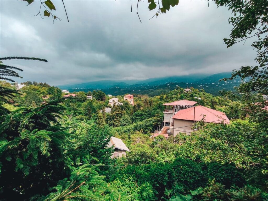 A view of Tikhisdziri a small village in Georgia with the red tiled roofs of the village houses lost in the greenery that surrounds the village. There are dark looming clouds and mountains looking purple in the distance. Off the beaten path Europe