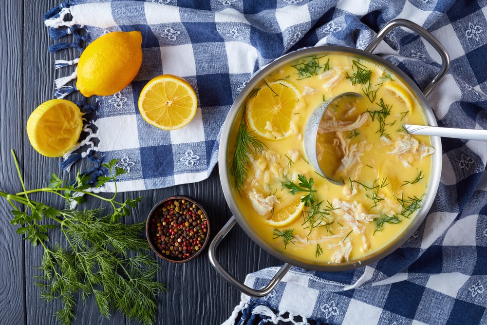 avgolemono - delicious creamy greek chicken soup with lemon, egg yolk, pasta risini and herbs in a casserole with soup ladle, on a wooden table with kitchen towel at the background, close-up