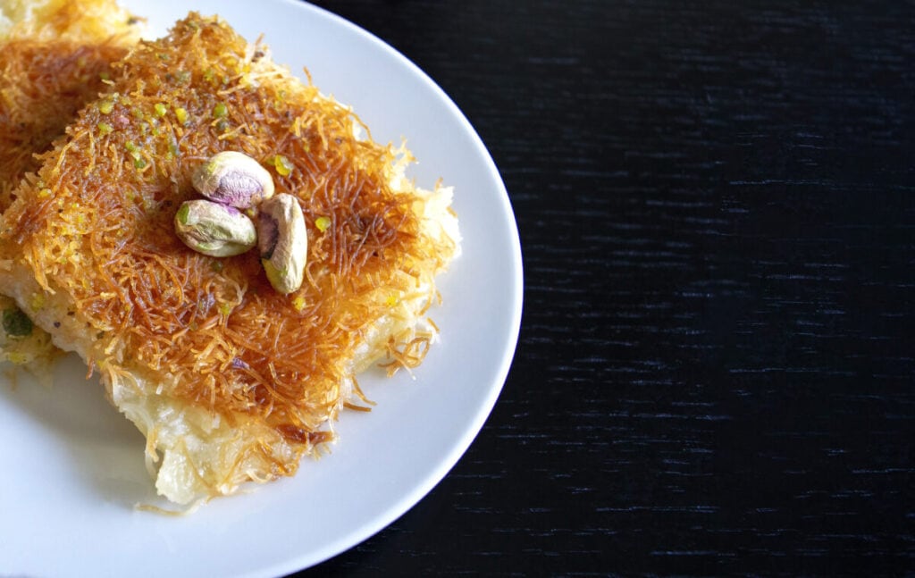 Traditional eastern dessert kunafa. Top view. Arabic cheese kunafa in white plate on dark wooden table with copy space. Traditional eastern dessert kunafa. Top view. Arabic cheese kunafa in white plate on dark wooden table with copy space. .