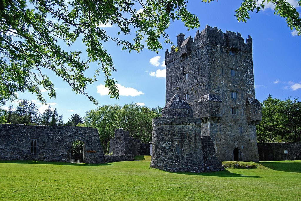 A ruined grey stone castle sits against a bright blue sky and green grass Aughnaure Castle in Connemara Ireland