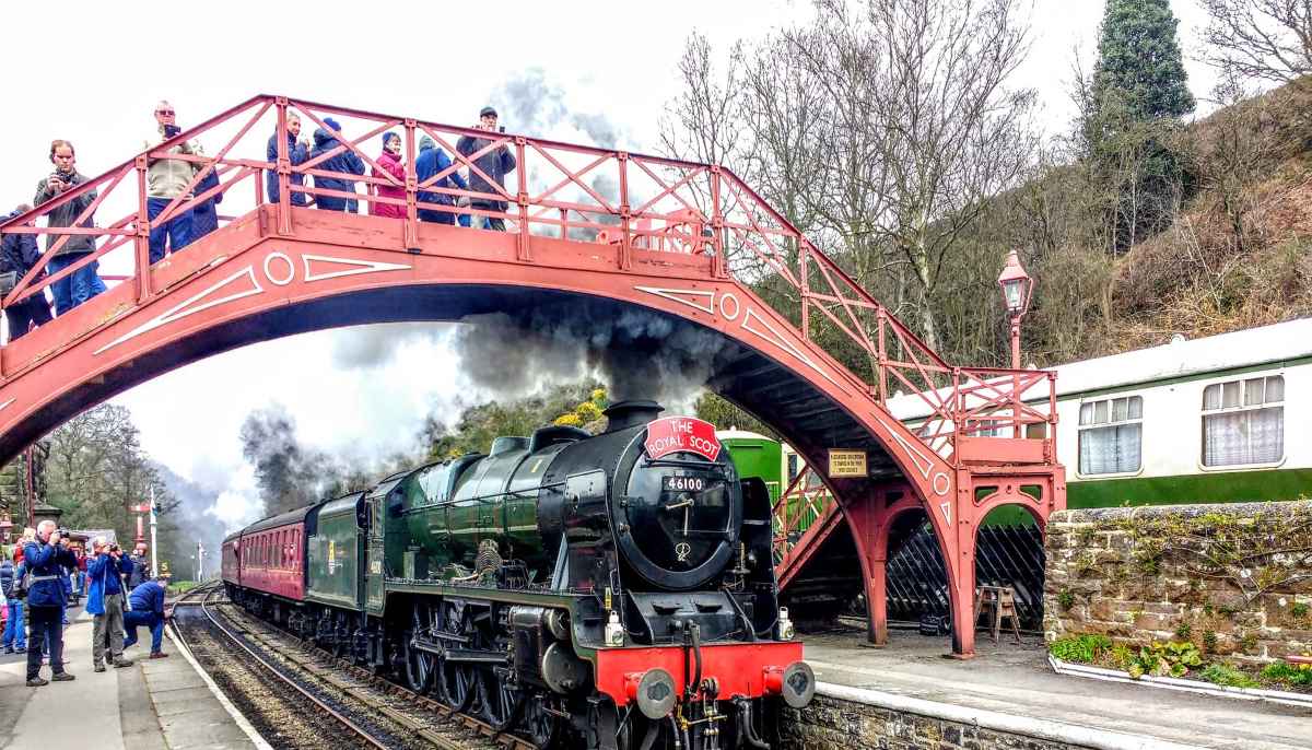 A steam locomotive passes under a red metal footbridge w people are standing and taking photographs.
