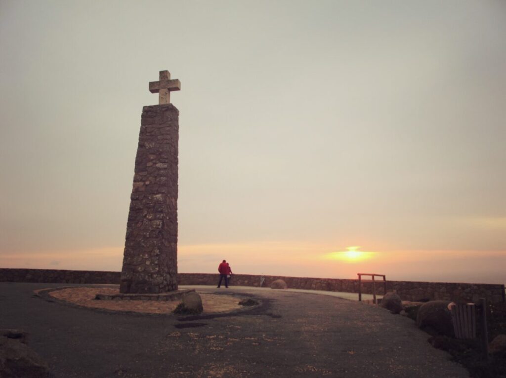 Cabo da Roca Portugal is the westernmost point of continental Europe and the best time to visit there is during sunset. There is a large stone monument with a cross at the top and a man viewing the sunset