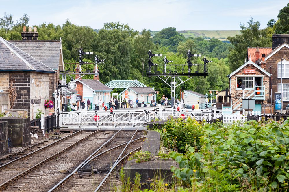 A railway crossing with white gates and signal gantries sits between stone buildings and a platform with people.