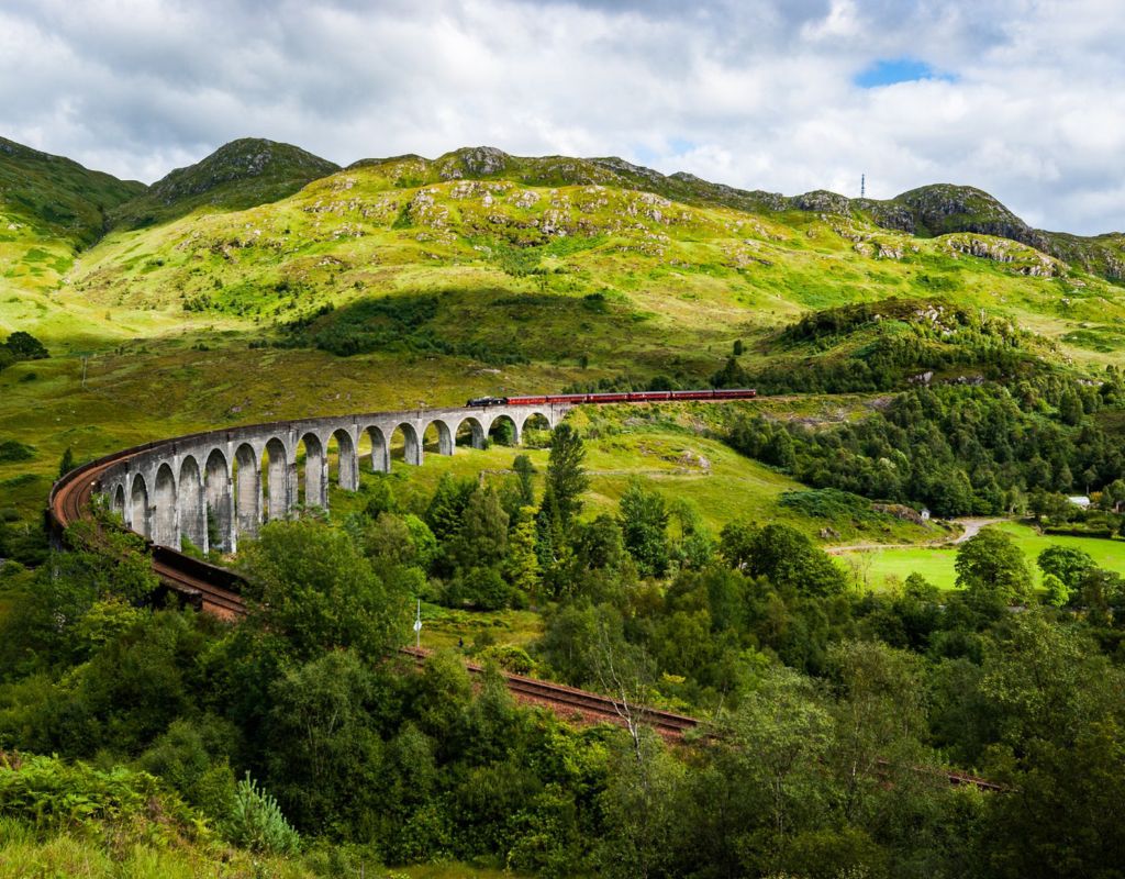 A red steam train travels across a long, curved stone viaduct set within a lush, hilly landscape.