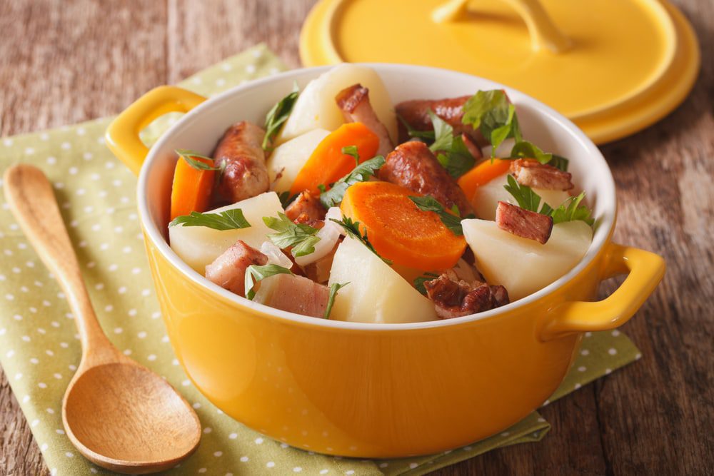 Traditional Irish dish is coddle with pork sausages, bacon and vegetables in a yellow pot on the table. A bright yellow Dutch oven filled with potatoes, and carrots and sausage with green cabble