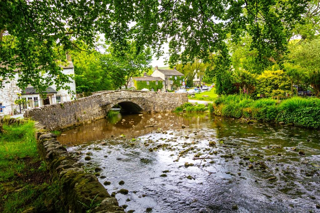 A stone bridge arches over a shallow stream near stone buildings surrounded by lush green trees. Malham Beck is a stream some 2 km long, running southwards through the valley beneath Malham Cove in the Yorkshire Dales, England