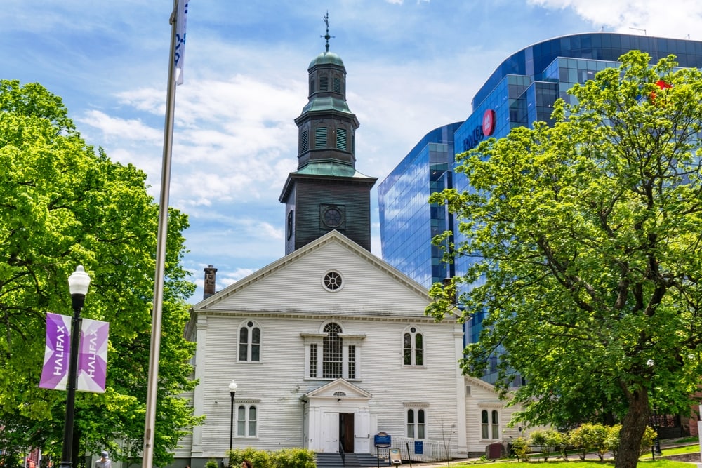 Historic St Paul's Anglican Church in Halifax haunted places in Canada. The church is a white clapboard building with a bell tower and georgian style gothic windows it is surrounded by trees and a Large GLass Bank of Montreal building sites behind it