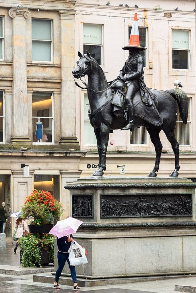 A coned statue of Duke of Wellington. It is raining. A woman with an umbrella walks next to the statue. There is a similarity between the coned statue and the woman with the umbrella.