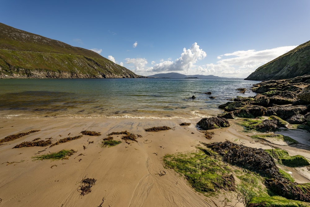 Hidden gems Ireland - Keem Beach a harbour of greenish blue water surrounded by rocky cliffs and a yellow sand beach with the tide bringing in seaweed to the rock pools