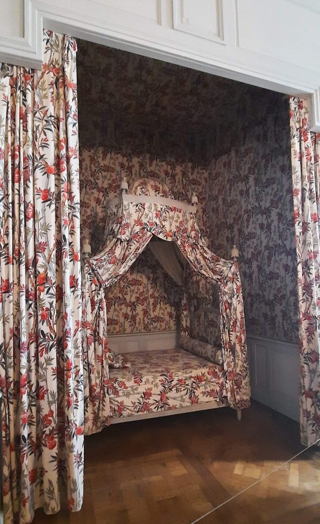 The Laurel bedroom with the bed tucked into a niche and decorated with a coronet of fabric. The curtains, walls and bed are all dressed in the same tiny floral print fabric