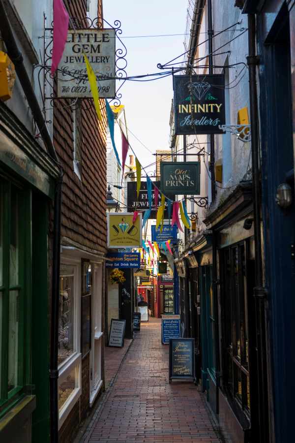 A narrow brick alleyway is lined with shop fronts and hanging signs, decorated with colorful bunting overhead.