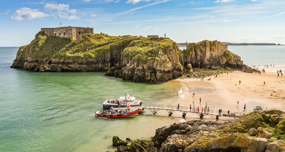 view of Castle Beach and St Catherine's Island in Tenby, West Wales at low tide. Visitors are getting off small cruise boats alongside a temporary jetty.