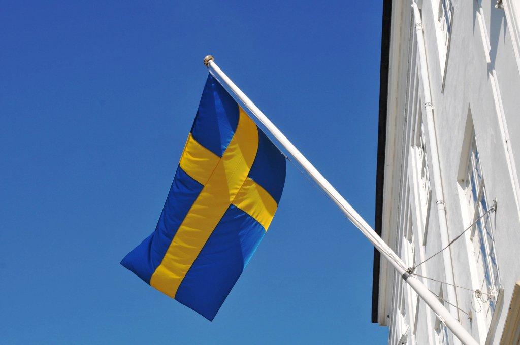 A Swedish flag flying from a white building. The flag had a back ground of blue with a bright yellow cross in the foreground