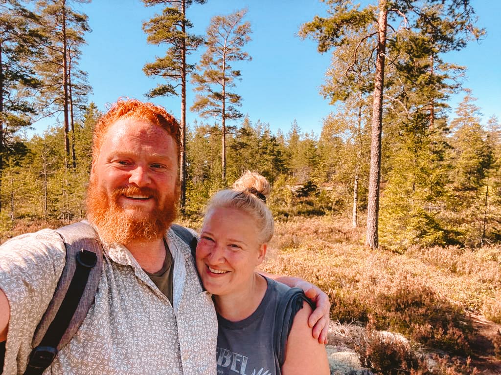 My husand and I in our forest land in Sweden. The young couple are surrounded by pine trees. He is red haired with a full beard and she is blond with her hair tied up in a pony tail