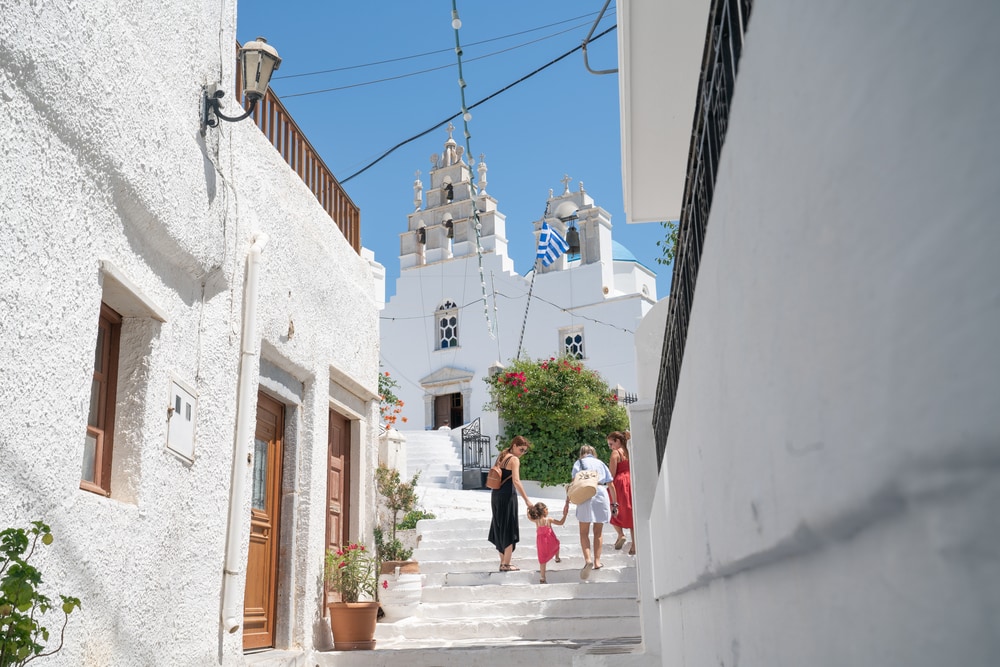 Three woman with small child walk up steps on path between white village buildings towards church in Greek Island village scene