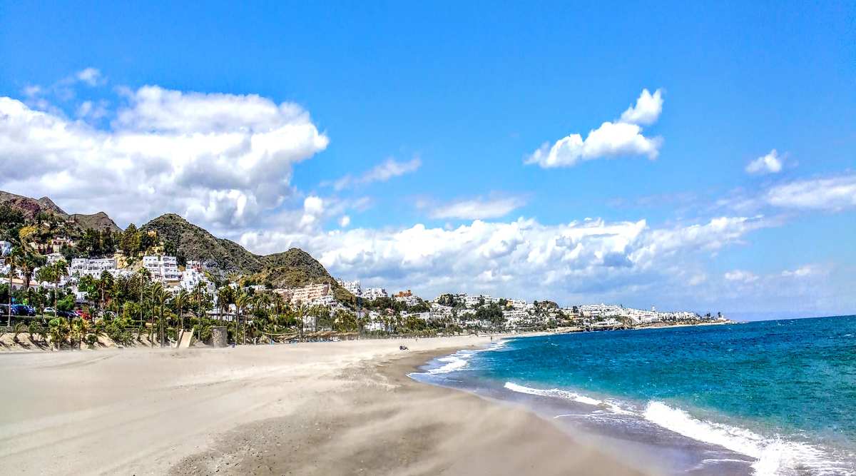 A sandy beach meets the blue sea below a hillside town with white buildings under a bright, partly cloudy sky.