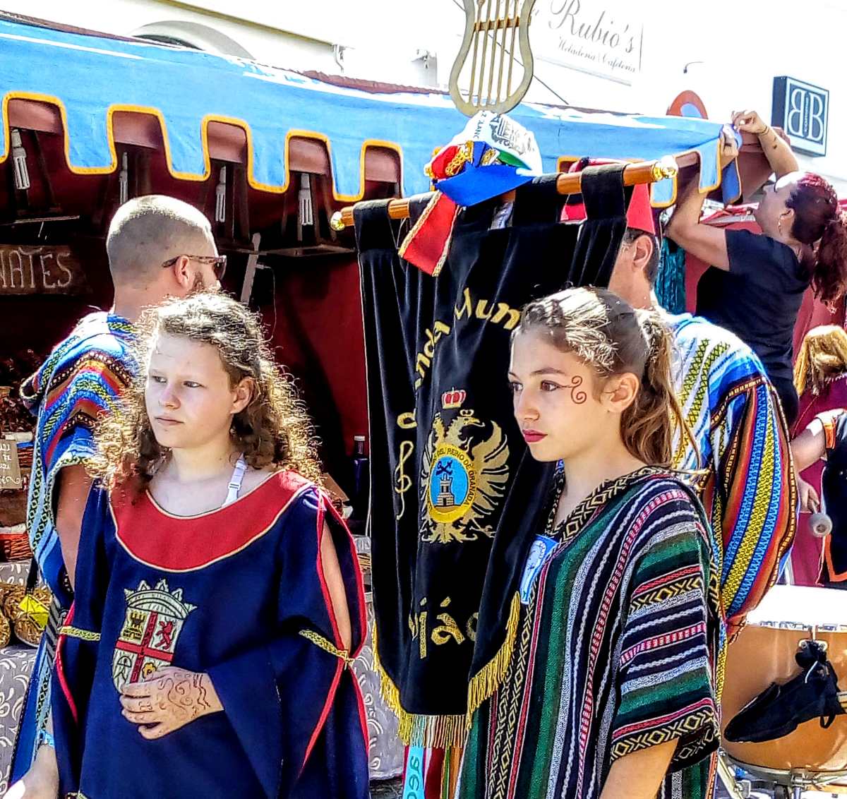 Two young people in historical-style clothing stand in front of a black banner with a gold emblem under a blue canopy.
