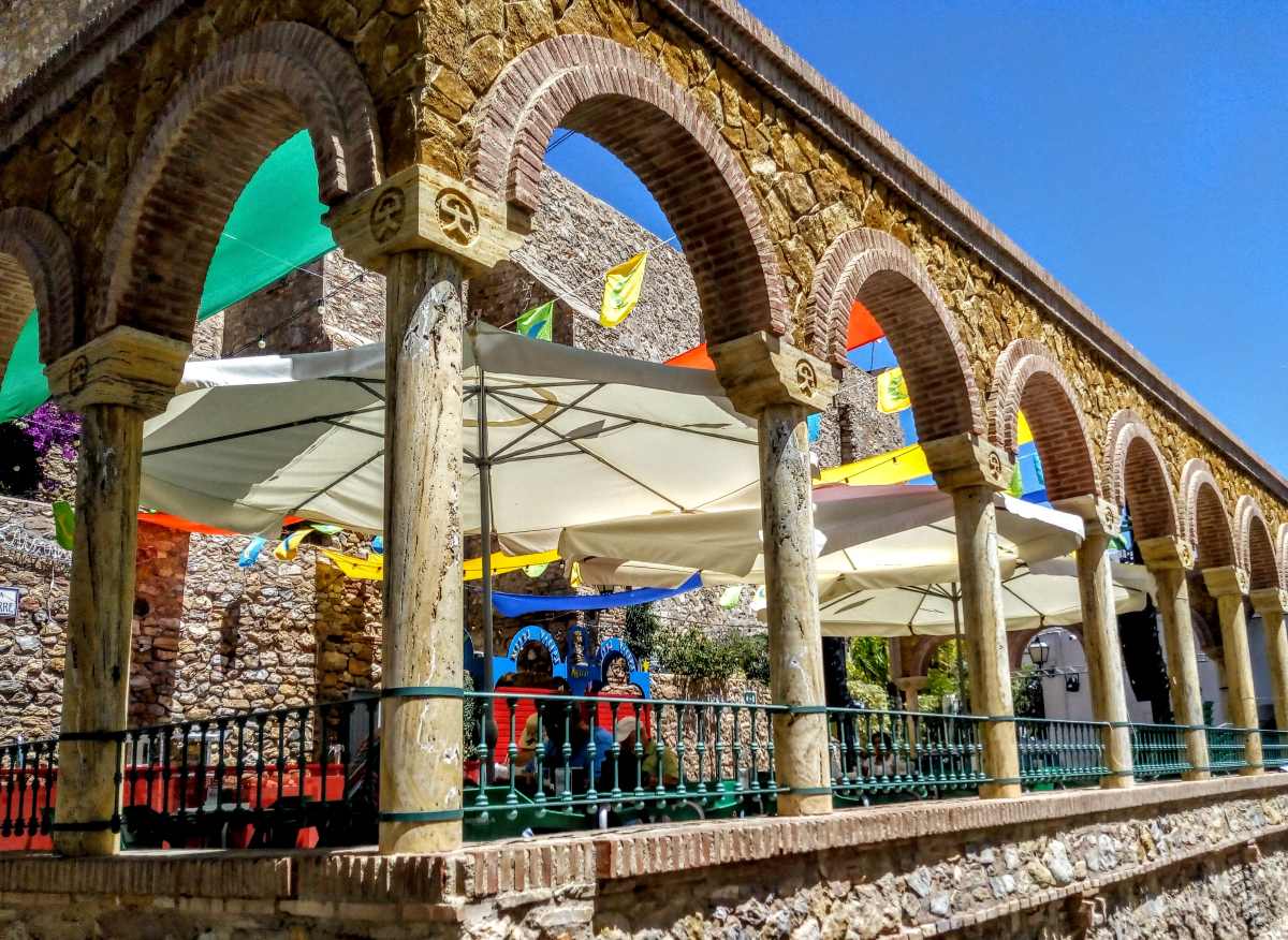 A stone arcade with arched openings overlooks a patio shaded by large white umbrellas and decorated with colorful flags.